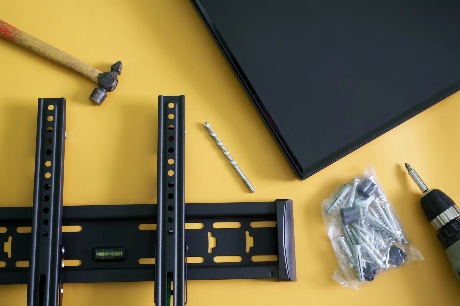 A tilting wall bracket with a built in level, a hammer, a masonry bit and a bag of screws and plugs on a yellow surface