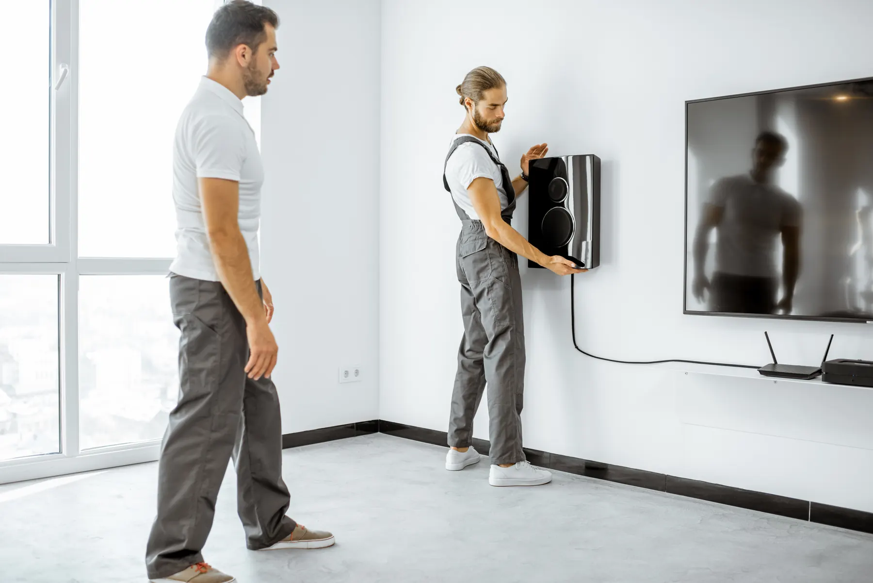 Two installers in an empty apartment, one fitting a small speaker beside an already mounted television