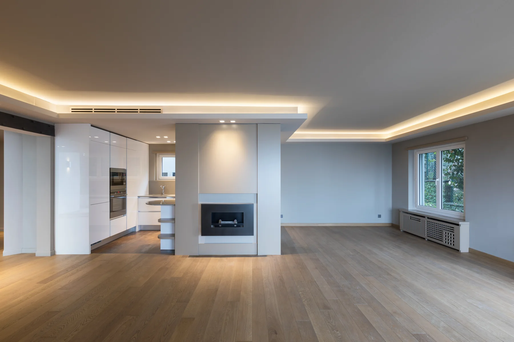 An empty open plan room with pale oak plank flooring, a white gloss kitchen at the far end, a recessed gas firebox set into a tall white partition wall, grey painted walls, cove lighting round the ceiling and a radiator under a window