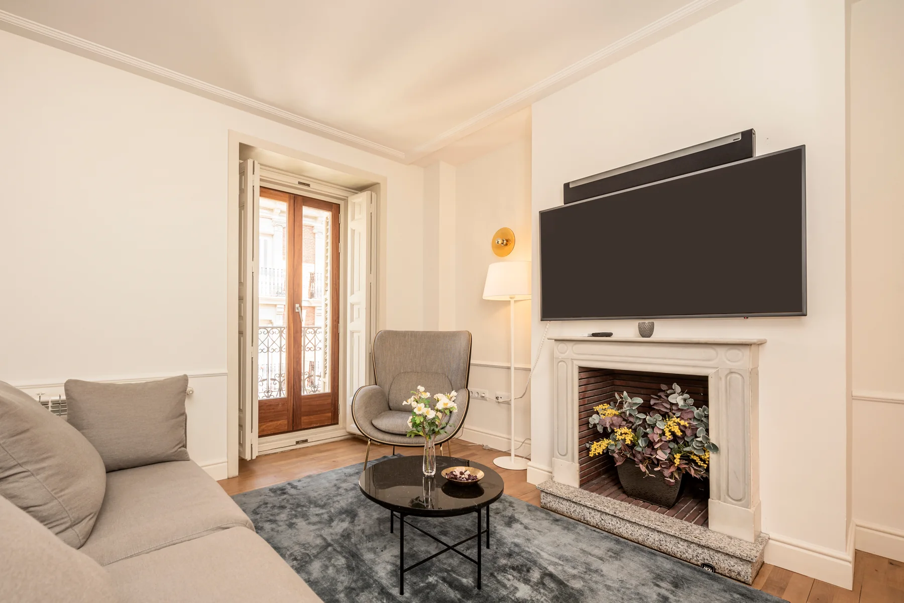 A wide black flat screen television, switched off, mounted on a cream wall above a carved white marble fireplace, with a black soundbar on the wall directly above the screen and dried flowers standing in the empty brick firebox