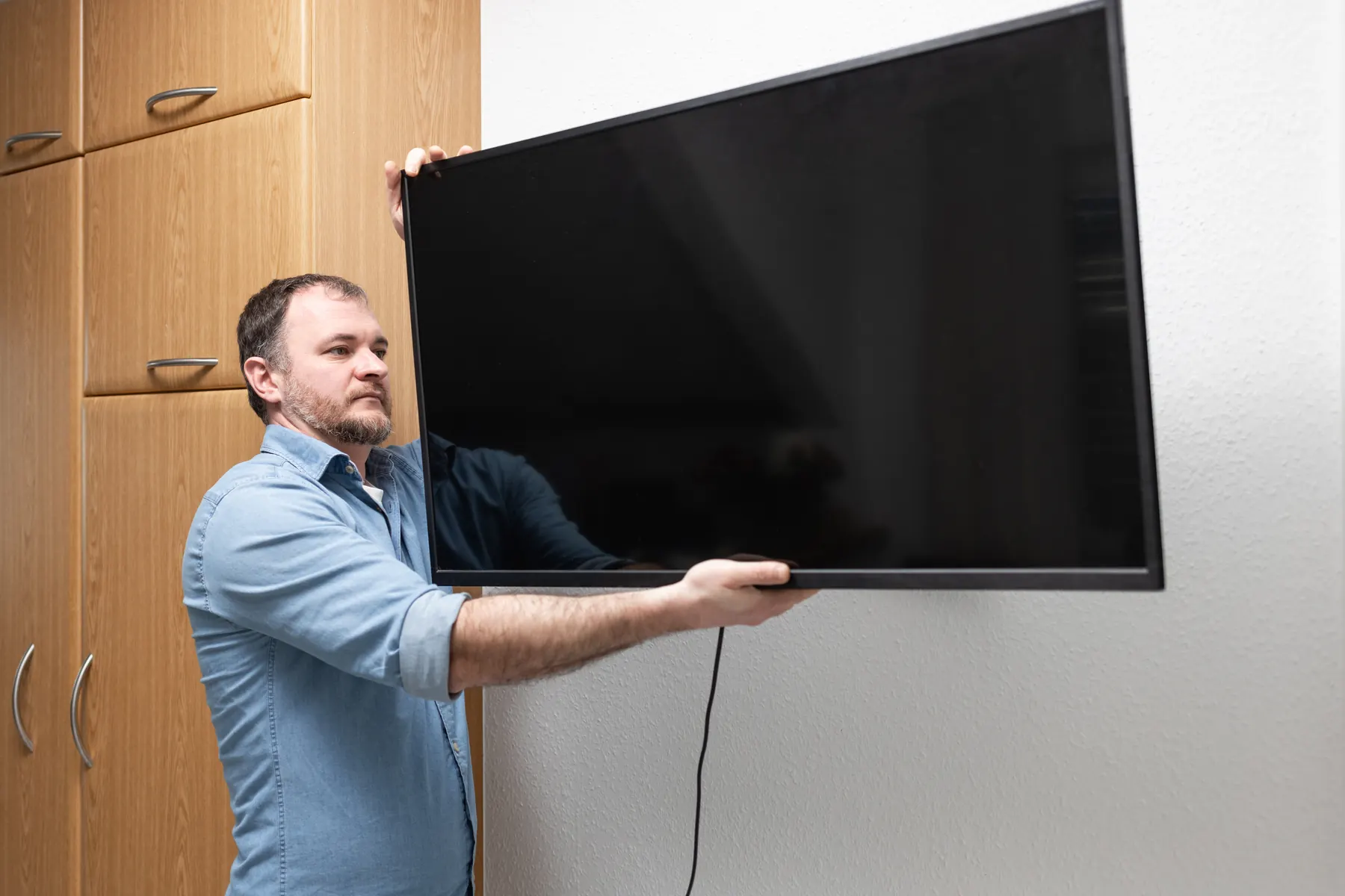 A man in a denim shirt holding a large television out from a white textured wall, ready to hang it