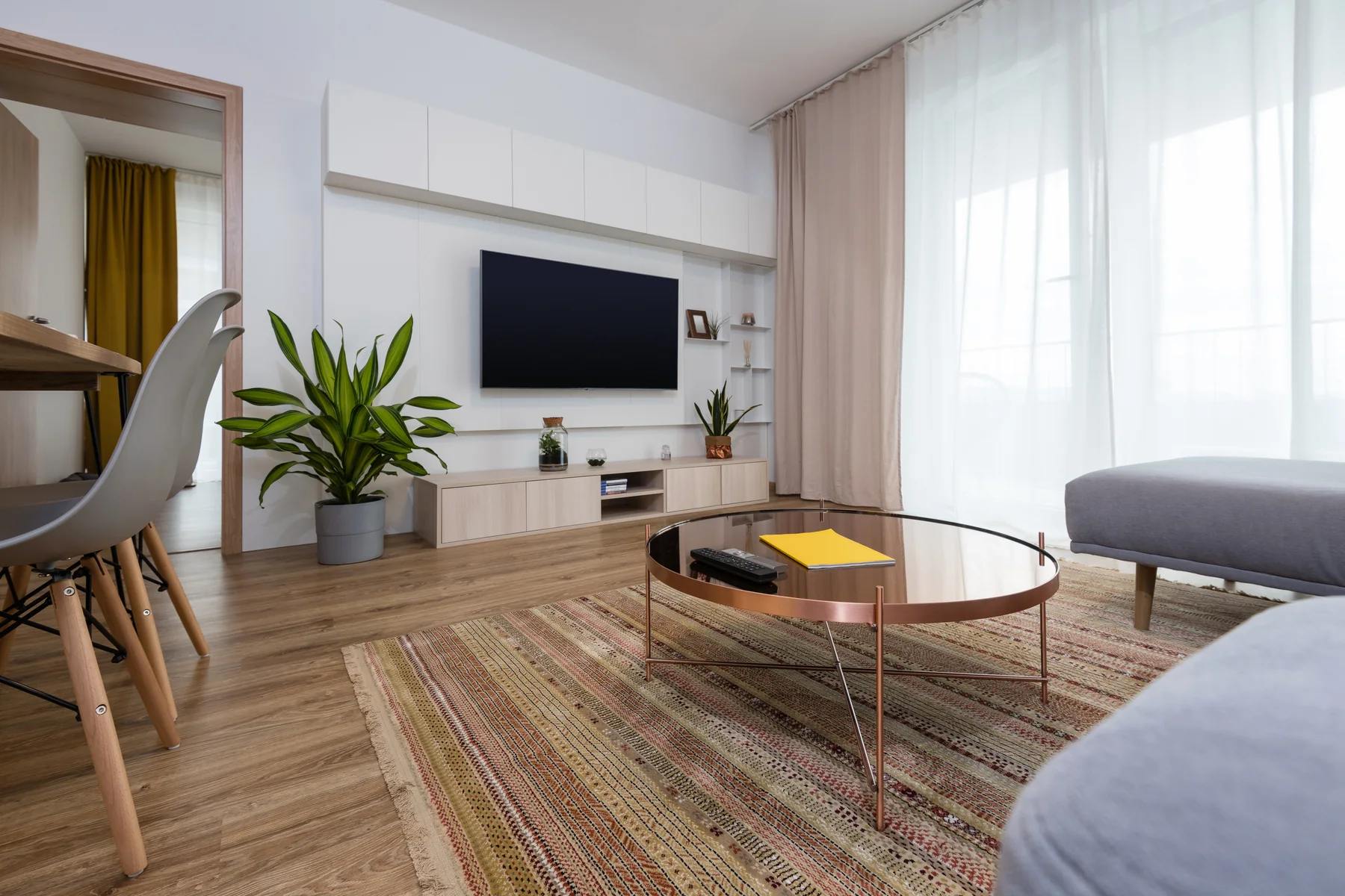 An angled daylight view of a living room with a black television, switched off, mounted flat on a white fitted wall unit above a pale oak sideboard, with a large green plant beside it, a copper and glass round coffee table on a striped rug and a grey sofa at the right edge