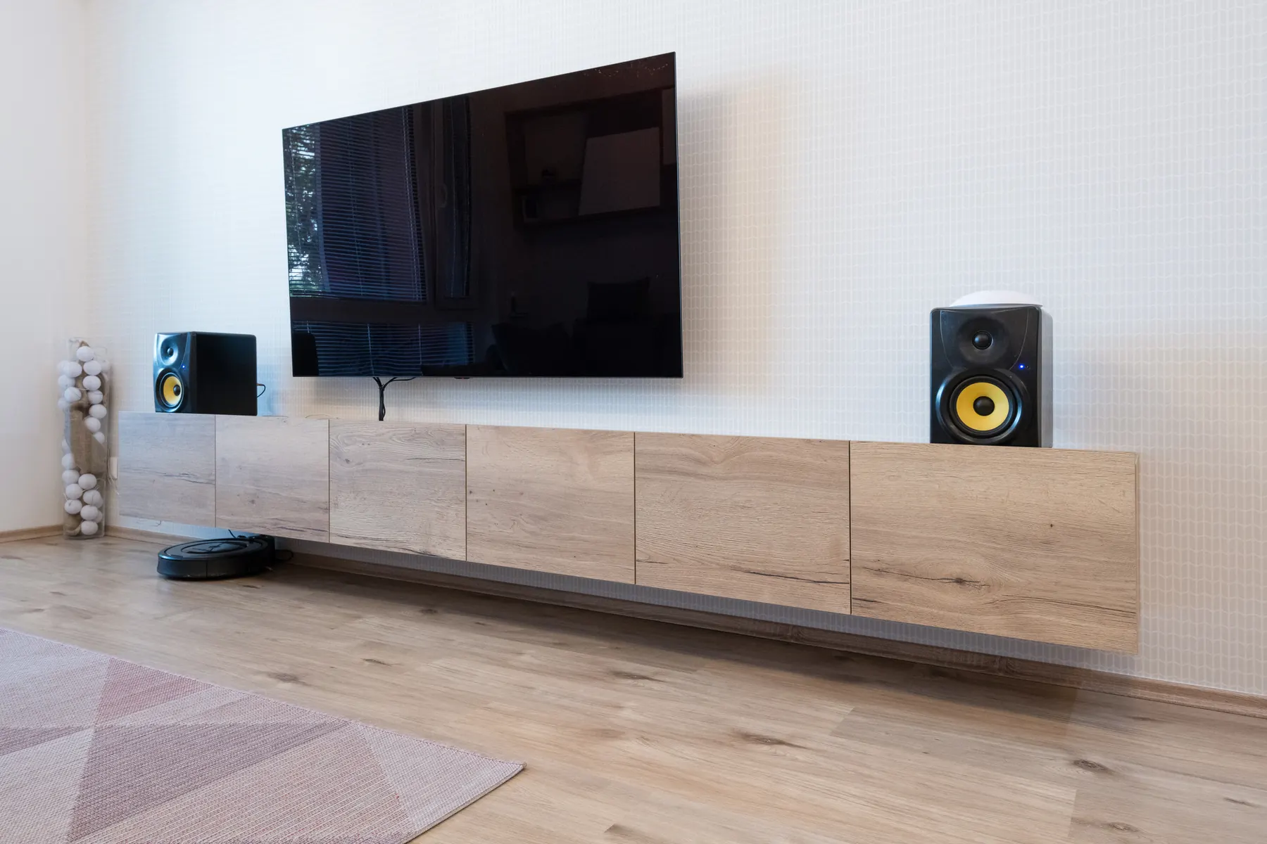 A black television mounted on a white textured wall above a long light oak sideboard holding two black bookshelf speakers with yellow cones