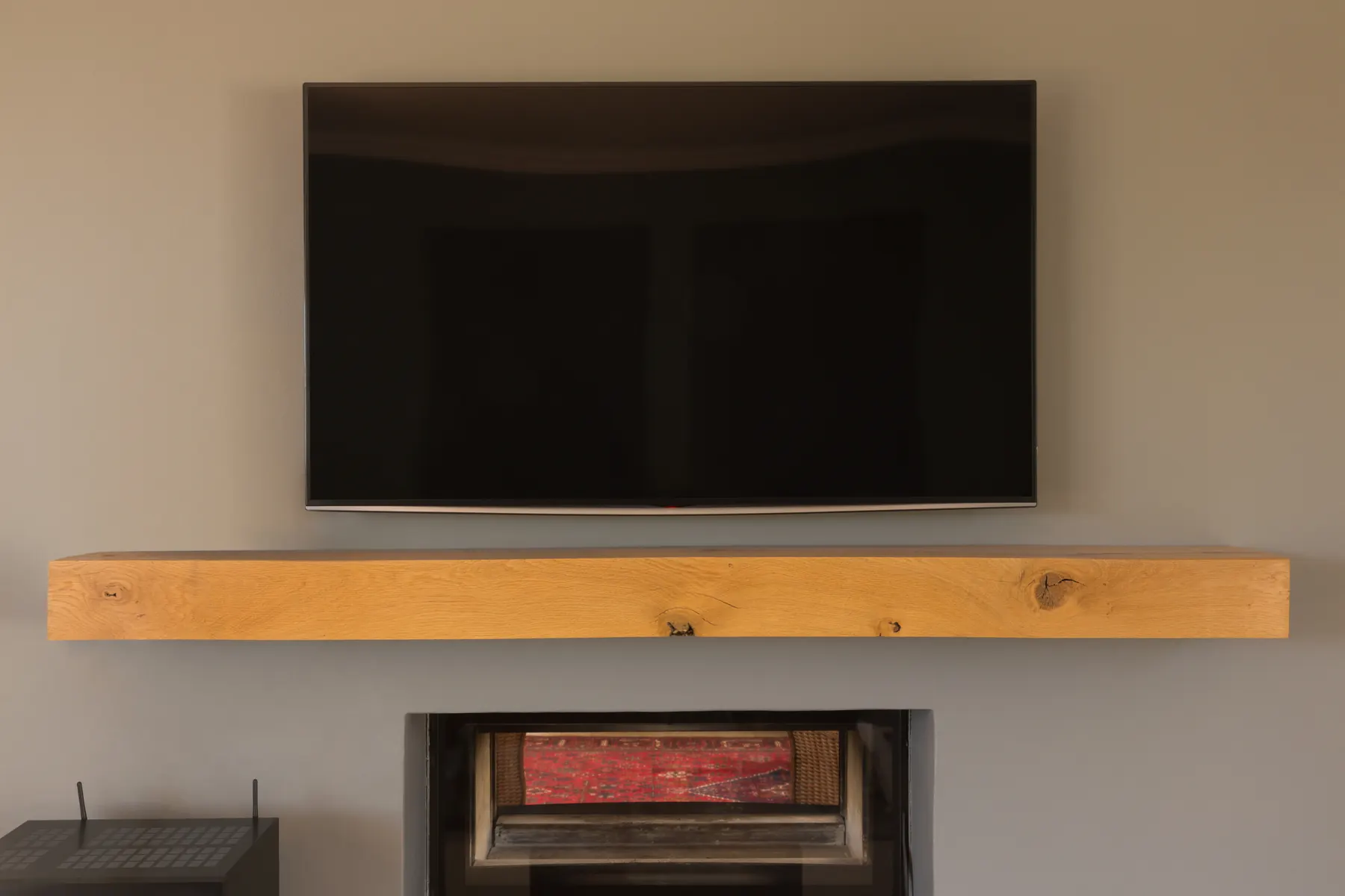 A television mounted on a grey beige wall above a thick oak mantel beam, with the dark metal top of a wood stove below it